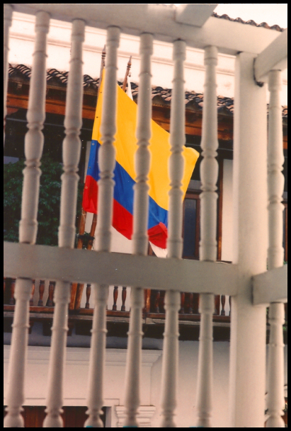 Colombia's flag with uneven color
            fields of yellow, blue and red seen through antique white
            fencepost rails