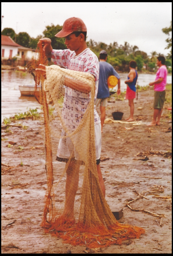 the teenage barber-fisherman
              straightening his net by the Rio Mojana in Santisima Cruz,
              with background of muddy shore, palms, people, houses