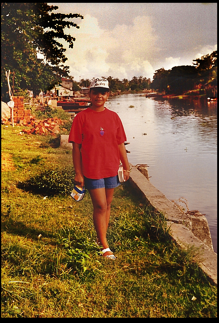 Sandi wearing neat
              loosely-fitting red t-shirt, stylishly faded jeans shorts,
              white Ducks Hockey cap, unscarred white sandals and a
              pleasantly serious smile, carrying a book in left hand and
              a box purse in right, on fluorescent green riverbank
              blessed by tropical rainy season sky and clouds reflected
              in rippling Rio