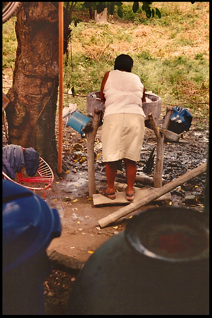 Victoria seen from back as
              she washes dishes in a big cement basin on criss-crossed
              logs in her back yard kitchen-'patio' surrounded by trees,
              grass and pots