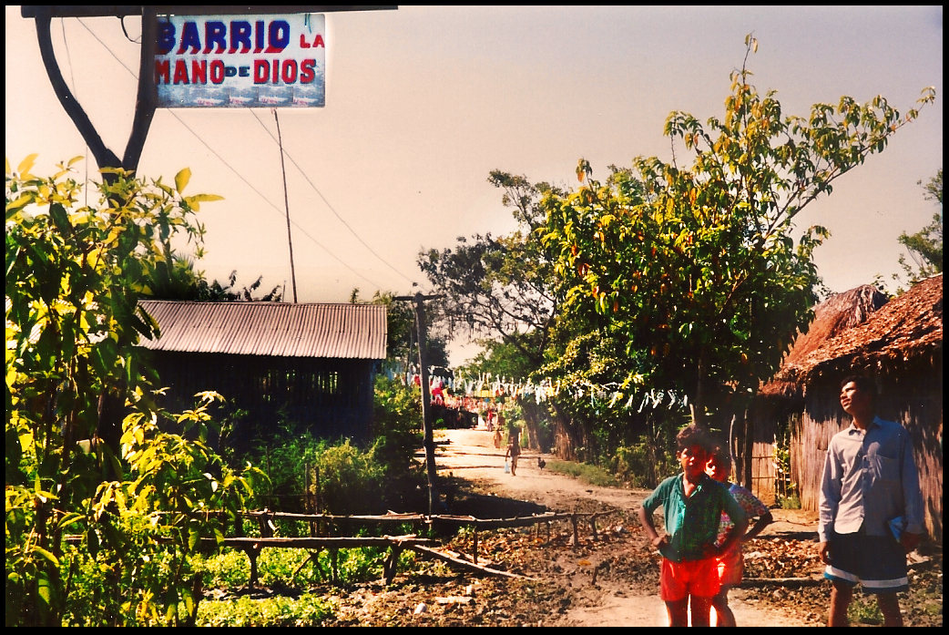 Pedro and 2
              young kids pose before thatch- and tin-roof neighborhood
              graced by a big welcoming sign high in the air: Barrio la
              Mano de Dios