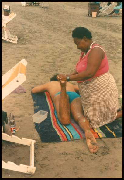 African-American woman massages Robbie's foot on
              beach towel at beach