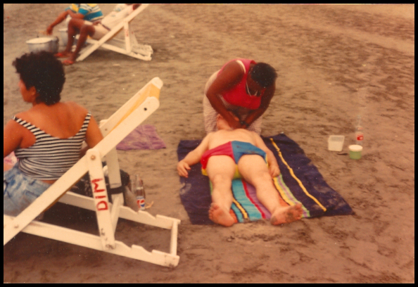 dark sandy
              becah, young woman in folding beach chair while much
              darker Afro-American woman works on the Dr.'s neck as he
              lies on back on multi-colored striped beach towel