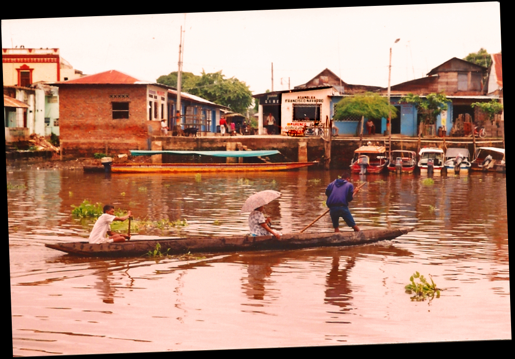 dugout canoe-taxi
              transporting lady with parasol passes Santisima Cruz
              waterfront shops and docked motor launches