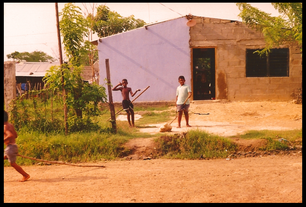 3 Pozón boys ride
              rudimentary stick horses in dirt street by cinderblock
              houses