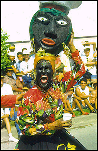 man face-painted and dressed as Jemimah-mammy (USA
            African slave woman) carries Africa-style a giant likeness
            of his own Black head ON HIS (own) (Black-painted) HEAD