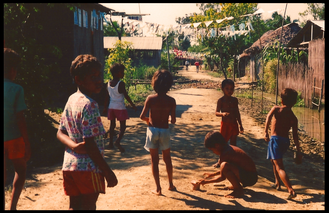 7 little barefoot boys in
            shorts play in dirt path amid thatch-roof shacks, flooded
            yards and fiesta street decorations in the Hand of God
            barrio