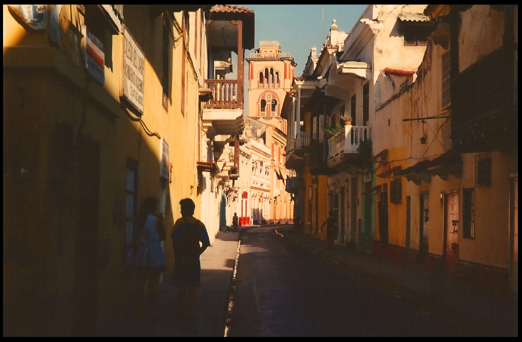 one-lane street lined with packed
            multi-colored houses and overhanging balconies, mostly in
            dark except upper stories and a tower at end