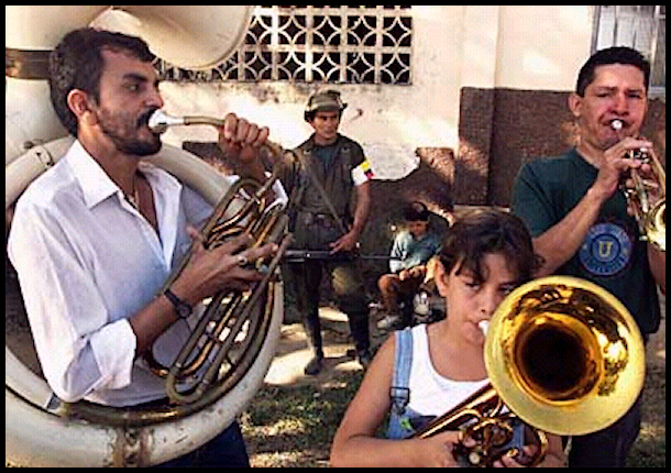 two local men on
            tuba and trumpet and a girl on baritone horn play while
            guerrilla in funky hat guards with automatic rifle