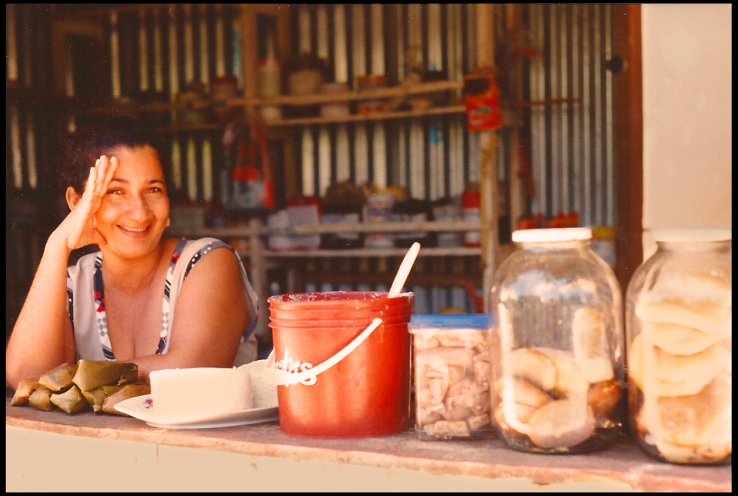 smiling
            middle-aged woman leans on shop counter flanked by home-made
            costeño snack foods in buckets and see-through jars