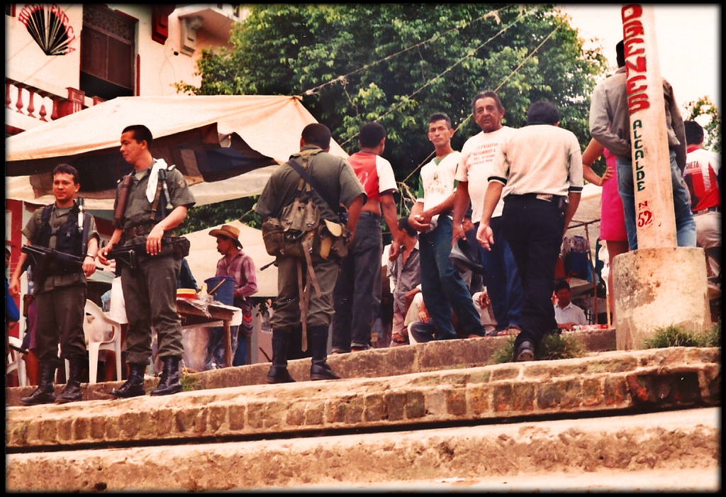 three government soldiers
              guard the top of the stone dock-step entrance to Santisima
              Cruz, busy market in plaza behind them