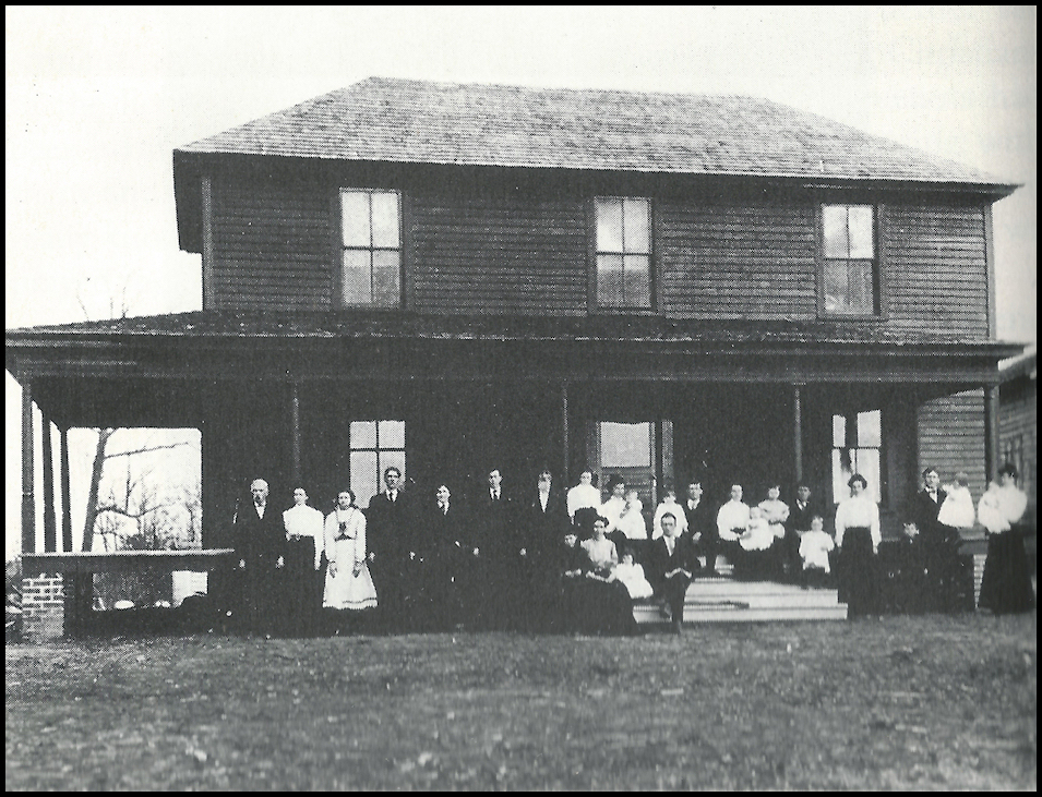 old black and
            white photo: roughly 1900 dark wood frame house with a large
            gathering of people in neat late-Victorian garb