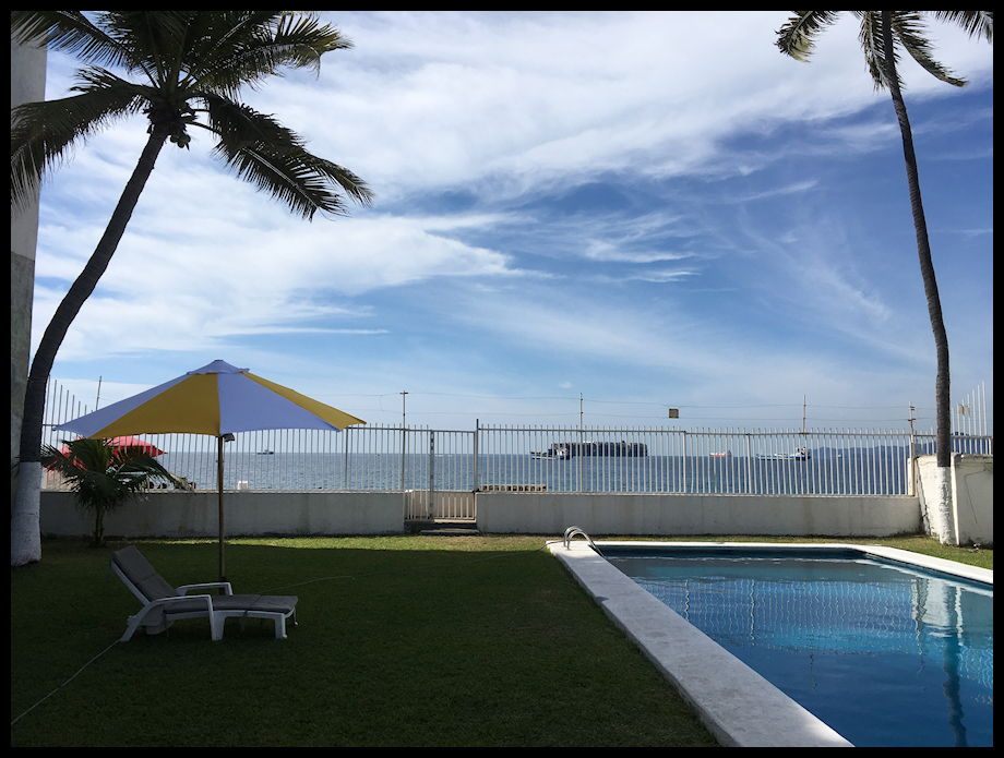 color: pool and lounge chair foreground;
                fence midground; bay with ocean-going vessels, including
                huge laden container ship; Mexican coastal mountains