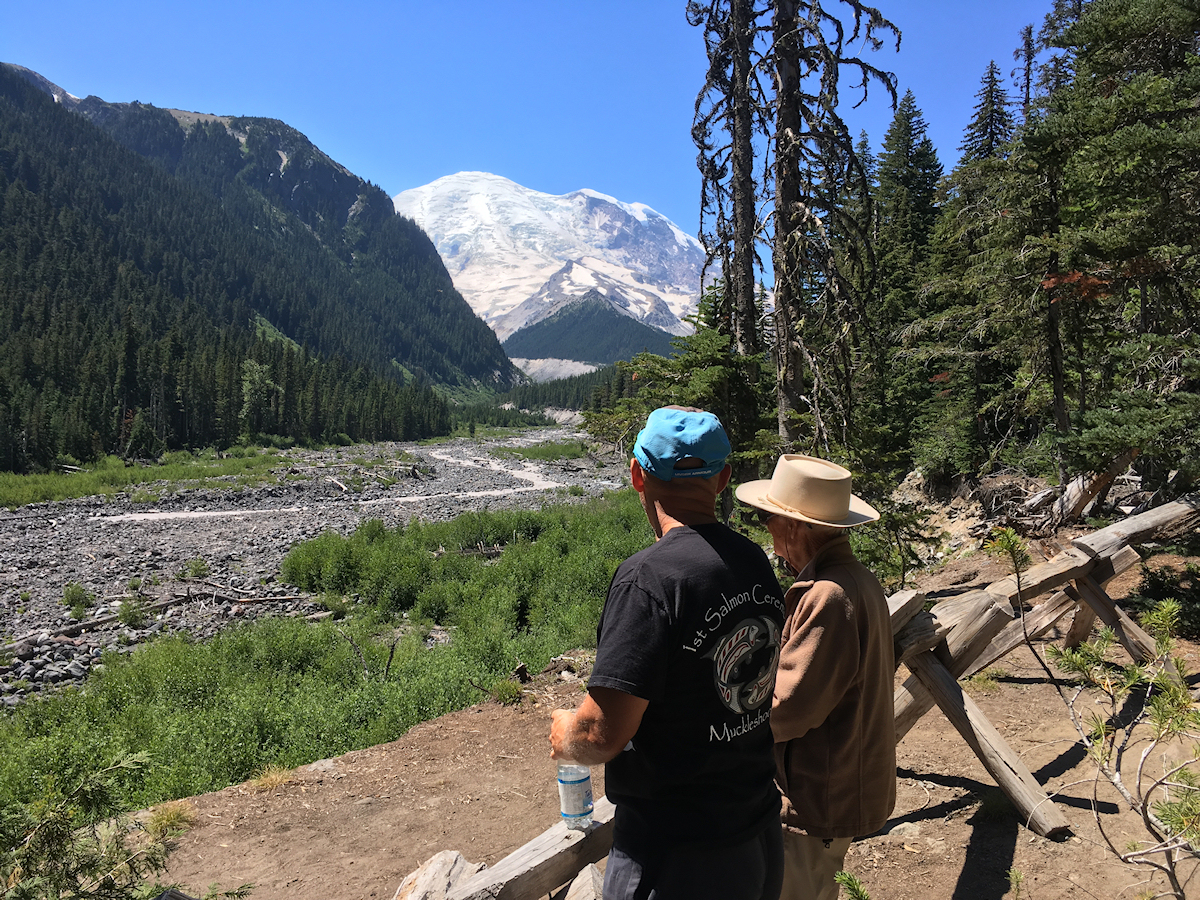 glacier-covered round peak, gorgeously
            clear blue sky, alpine river, fir forest and two men
            admiring the scene