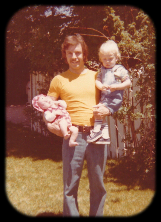 color photo, old and almost
              blurry, 30-ish man standing in yard cradling sleeping
              infant baby girl in right arm while 2-year-old boy sits on
              his left forearm