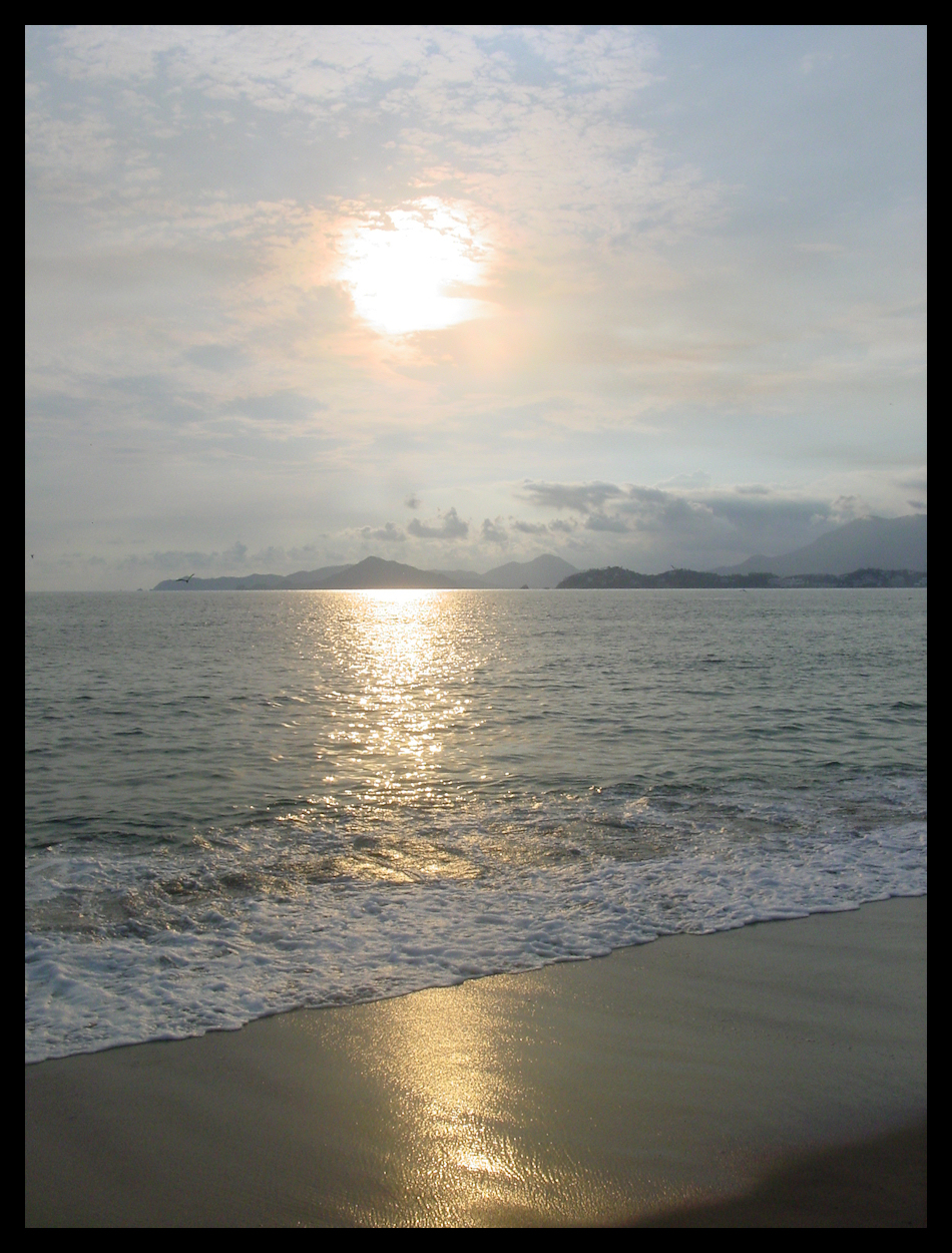 burning hot globe partly
            covered by late-day clouds, hangs over bay, mountains in
            distance, beach and waves in foreground